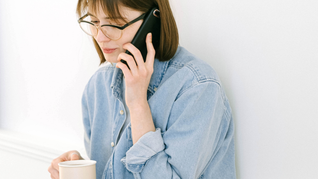 壁にもたれかかり、マグカップを持ちながら電話をしている女性の様子